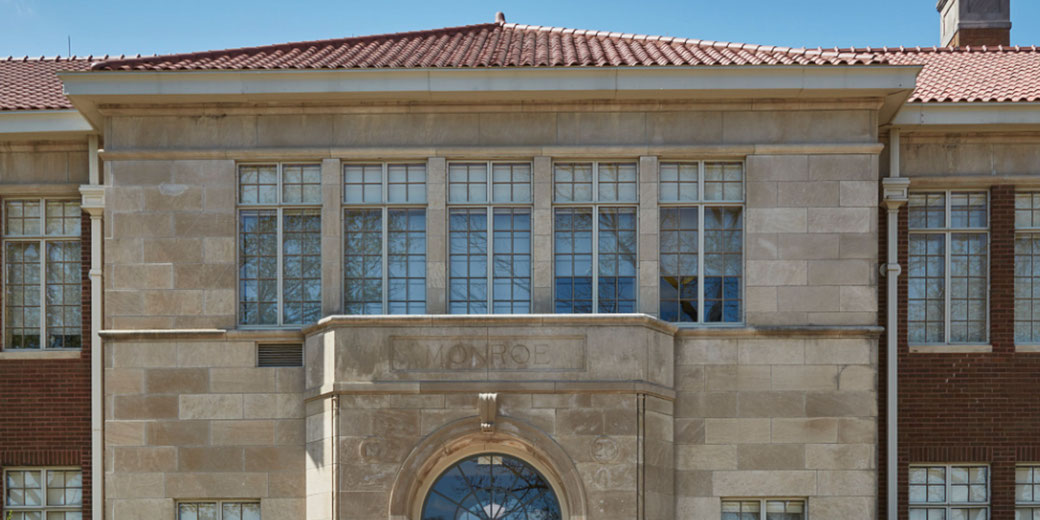 A historic stone building with large grid windows and a red tile roof. "Monroe" is engraved above the arched entrance.