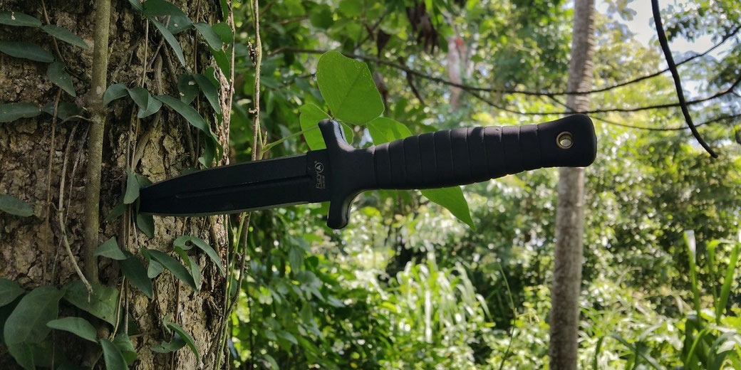 Black tactical knife embedded in a tree trunk surrounded by dense green foliage and vines in a forested area.