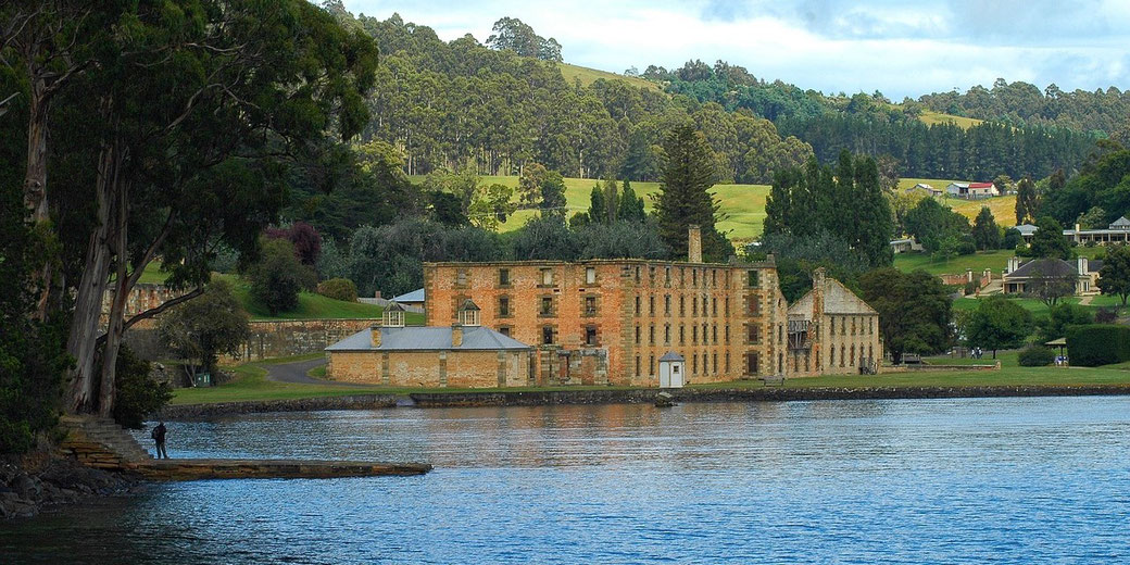 A large historic brick building stands by the water’s edge, surrounded by trees, grassy hills, and scattered houses in the distance.