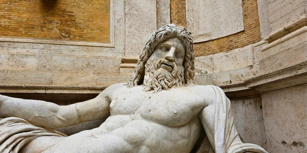 Stone statue of a reclining bearded male figure with muscular torso, set against a curved brick and stone wall, showing age marks and sculpted detail.