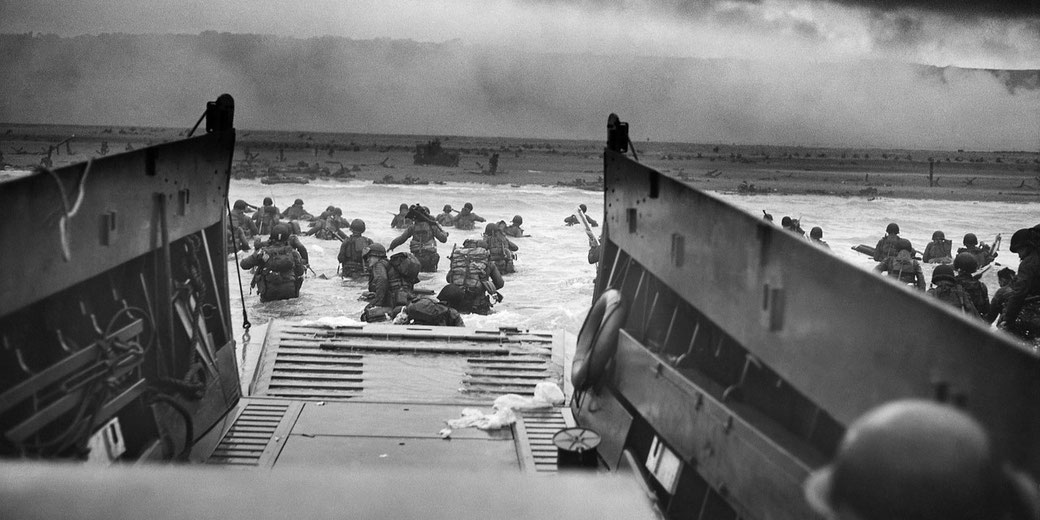 Soldiers wade through water from a landing craft toward a heavily fortified beach under smoke-filled skies during a military assault.