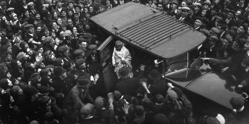 Mahatma Gandhi exits a car, greeted by a dense crowd of admirers and watched over by police in uniform.