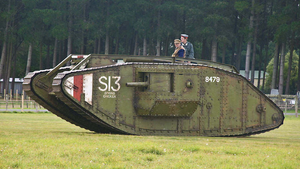 A restored WWI British Mark IV tank named "Spring Chicken" with two people on top, driving across a grassy field near a forest.