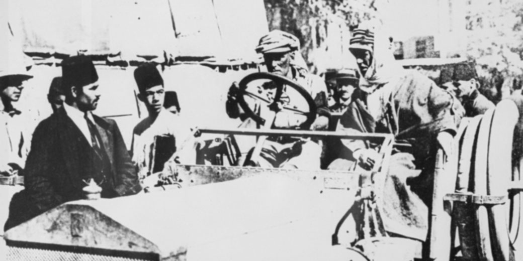 A group of men in traditional Middle Eastern and military attire gather around an old vehicle, with one man seated at the wheel, suggesting a moment during a historical wartime encounter.
