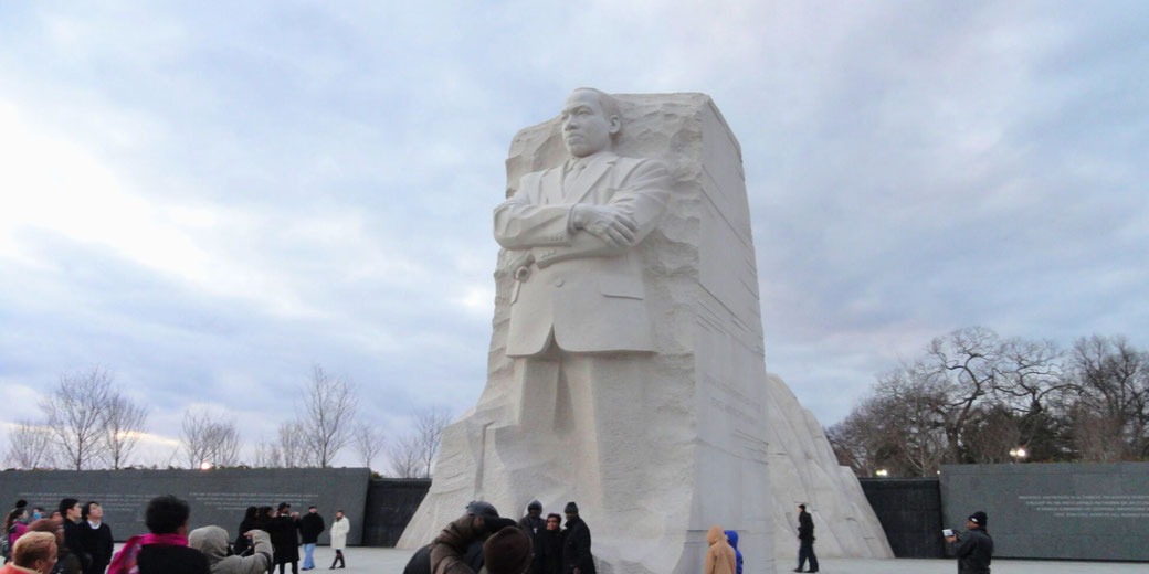 The Martin Luther King Jr. Memorial in Washington, D.C., featuring a towering stone sculpture of Dr. King with arms crossed, surrounded by visitors under a cloudy sky.
