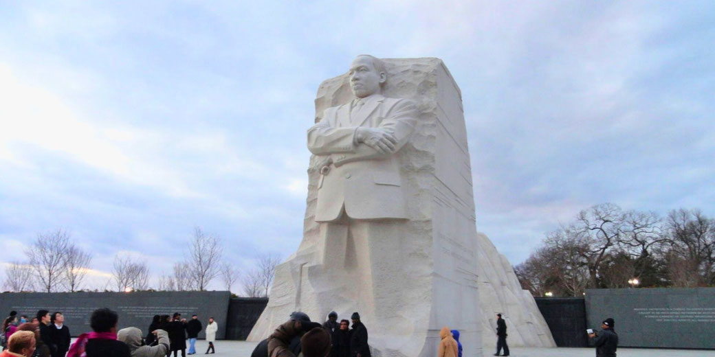 The Martin Luther King Jr. Memorial in Washington, D.C., featuring a towering stone sculpture of Dr. King with arms crossed, surrounded by visitors under a cloudy sky.
