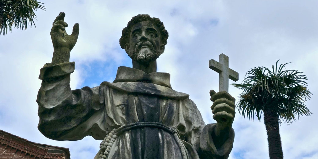 A weathered stone statue of a Christian saint or monk holding a cross in one hand and raising the other. The figure wears a robe with a rope belt. Palm trees and a cloudy sky are behind.