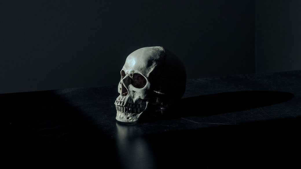 A human skull placed on a dark surface, partially illuminated by a beam of light, creating a dramatic and somber atmosphere with deep shadows surrounding it.