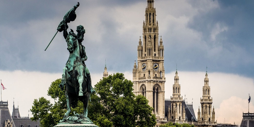 An equestrian statue with a raised flag stands before a tall clock tower and ornate buildings under a cloudy sky.
