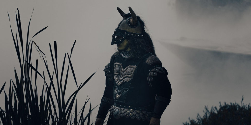 A person in horned armor and a dark mask stands near tall grass by a foggy body of water.
