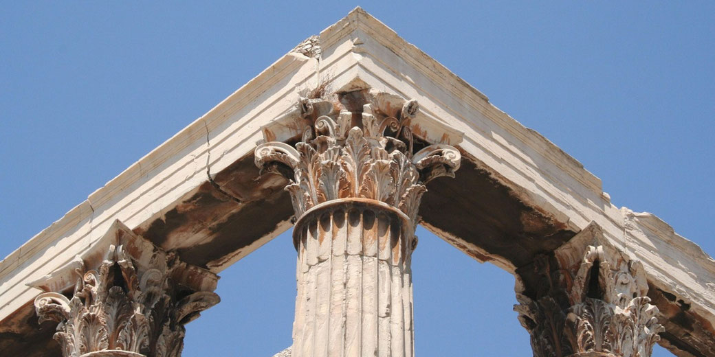 Close-up of ancient Corinthian columns supporting a weathered marble pediment, showcasing intricate acanthus leaf carvings under a clear blue sky.