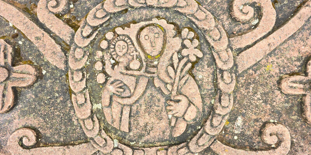 A stone carving depicting Mary holding baby Jesus, encircled by an ornate border. The artwork has a stylized, medieval or folk art appearance, with intricate detailing and weathered texture.