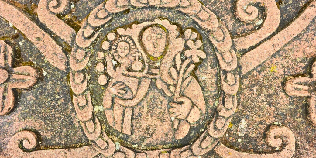 A stone carving depicting Mary holding baby Jesus, encircled by an ornate border. The artwork has a stylized, medieval or folk art appearance, with intricate detailing and weathered texture.