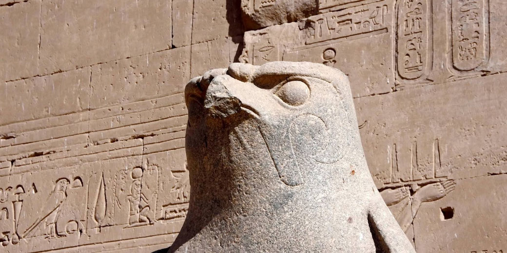 Close view of a carved falcon statue before an ancient Egyptian temple wall with hieroglyphs and stone textures in warm daylight.