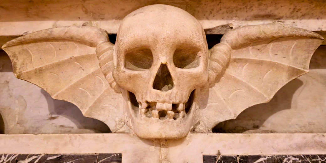 Stone carving of a human skull with bat-like wings extending from each side, set into a wall or tomb structure.