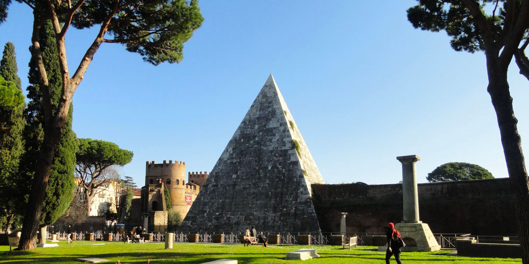 Large stone pyramid stands among trees and ruins under a clear blue sky, with people walking and sitting on benches nearby.