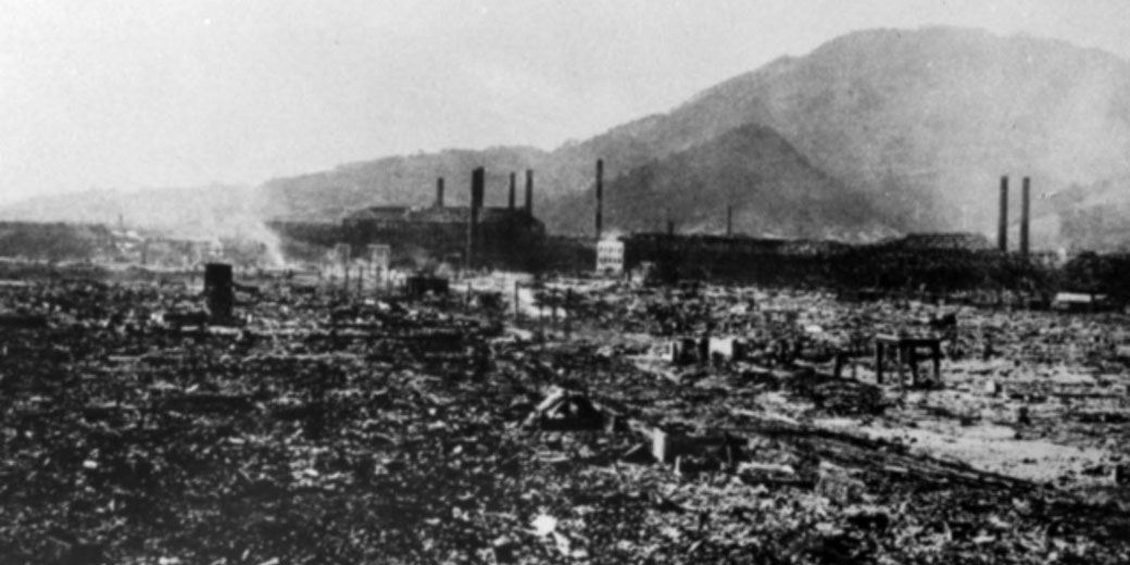 A black-and-white image shows the scorched ruins of Nagasaki after the atomic bombing, with smoke rising and factory chimneys in the background.