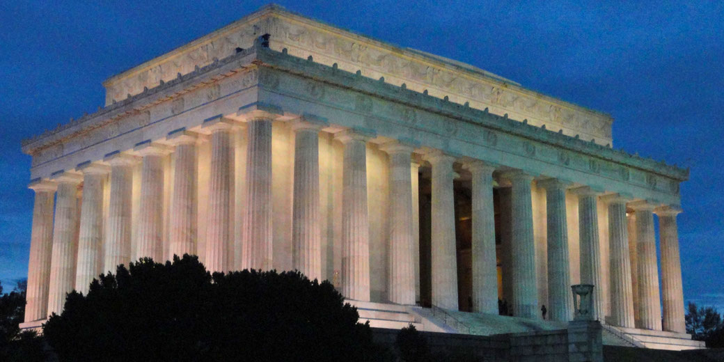 Lincoln Memorial at dusk, illuminated with soft lighting. The iconic neoclassical structure features large columns and a detailed frieze, set against a deep blue evening sky.