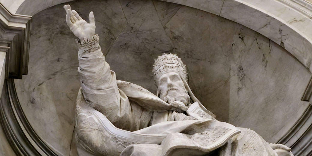 A marble statue of a pope in St. Peter's Basilica. The figure wears a papal tiara and ornate robes, raising one hand in a gesture of blessing.