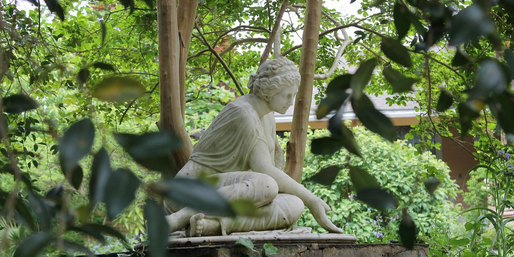 A marble statue of a seated figure is nestled in a lush garden, partially obscured by leaves. 