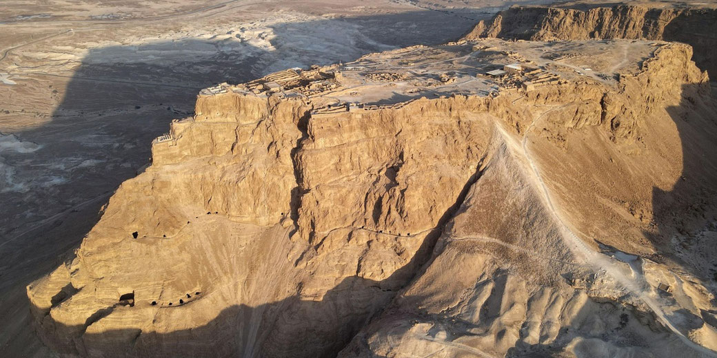 Aerial view of Masada, an ancient fortress on a desert plateau with rugged cliffs and archaeological ruins.
