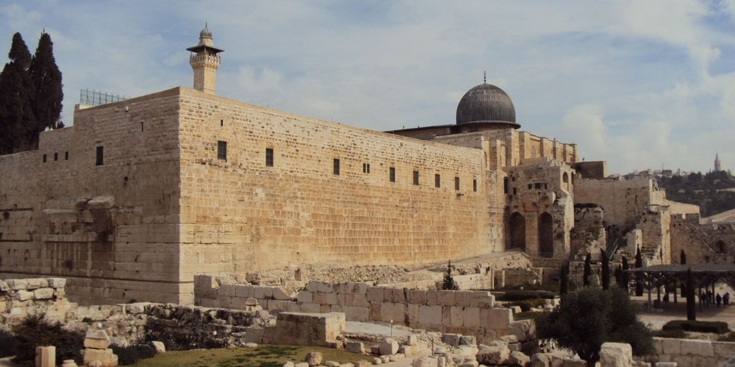 A large stone building with a black dome and minaret rises above ancient ruins and pathways under a partly cloudy sky.