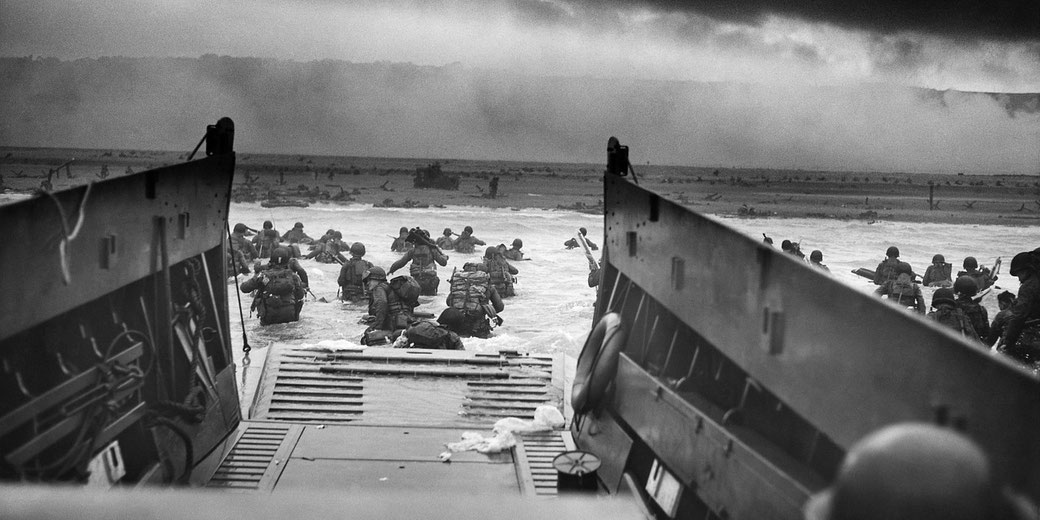 Soldiers disembark from a landing craft and wade toward a fortified beach during a wartime amphibious assault.