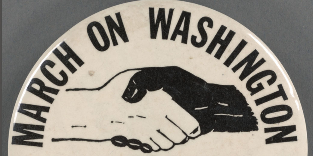 White circular button reads “March on Washington” arched above a black hand and a white hand clasped in a handshake representing unity.