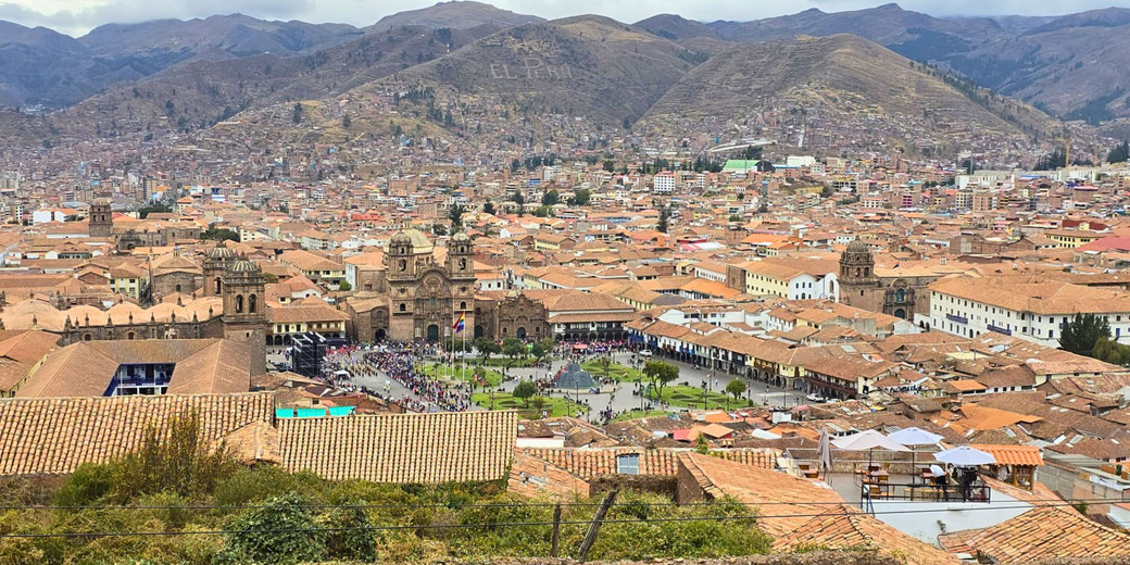 A panoramic view of Cusco, Peru, showcasing Plaza de Armas, colonial architecture, and terracotta rooftops. The surrounding mountains feature "Viva El Perú" carved into the hillside.