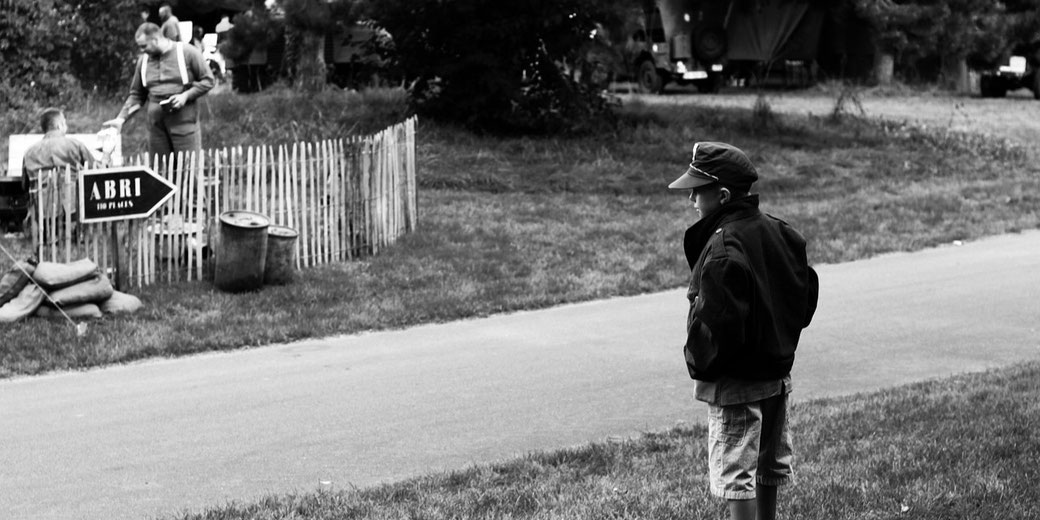 A boy in oversized military-style clothing stands alone by a road, watching reenactors in uniform near a sign reading “ABRI” in a black-and-white wartime-themed scene.