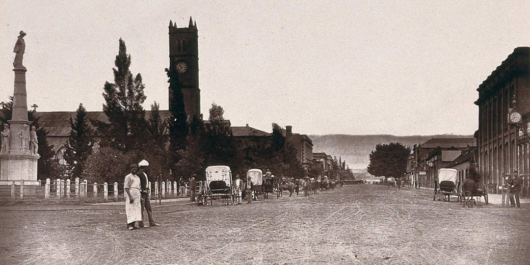 Historic street scene with horse carriages, a clock tower, and pedestrians on a wide dirt road in an early town setting.