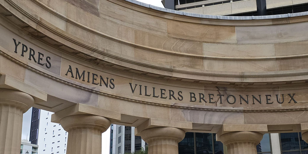 Curved stone memorial wall with engraved names of World War I battle sites: Ypres, Amiens, Villers Bretonneux, supported by columns.