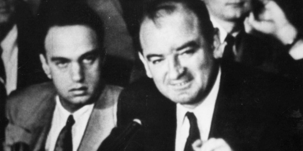 Black and white photo of two men in suits seated close together during a tense public hearing or meeting, with microphones and other people blurred in the background.