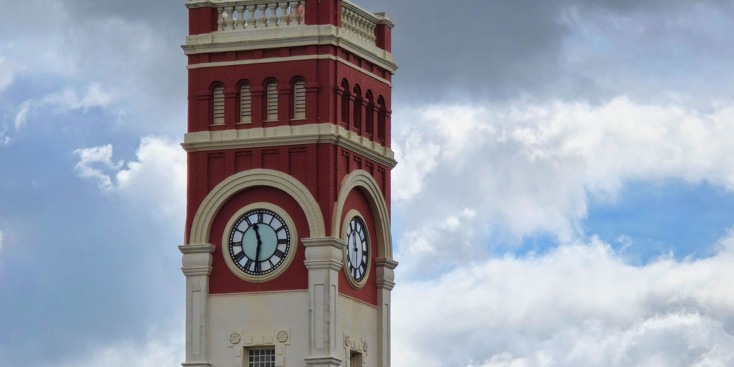 A red and white clock tower with large round clocks on each side stands against a cloudy sky with patches of blue visible. Its architectural style features ornate details and arched windows.