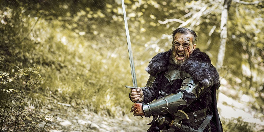 A battle-hardened warrior in fur-trimmed armor stands in a forest, gripping a sword with bloodied hands.