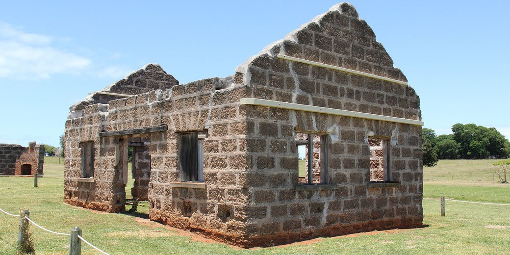 A weathered stone building ruin stands in an open grassy field under a bright blue sky. The structure has missing windows, doorways, and no roof, with remnants of wooden supports on its walls.