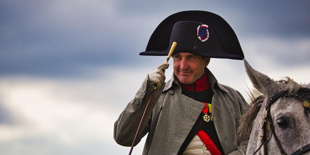A man in Napoleonic uniform tips his hat while mounted on a horse, smiling with a cloudy sky behind him.