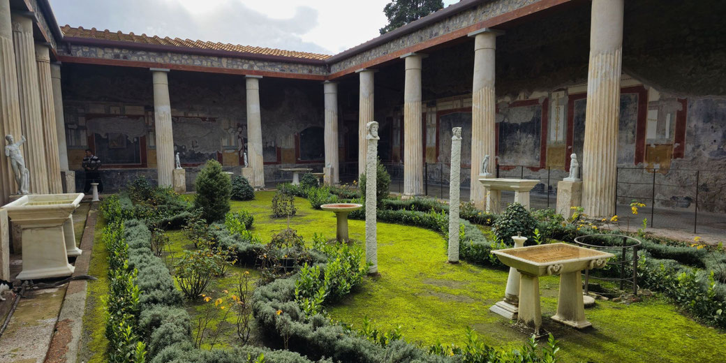 The garden of a Roman villa, featuring lush greenery, classical statues, fluted columns, and decorative fountains.