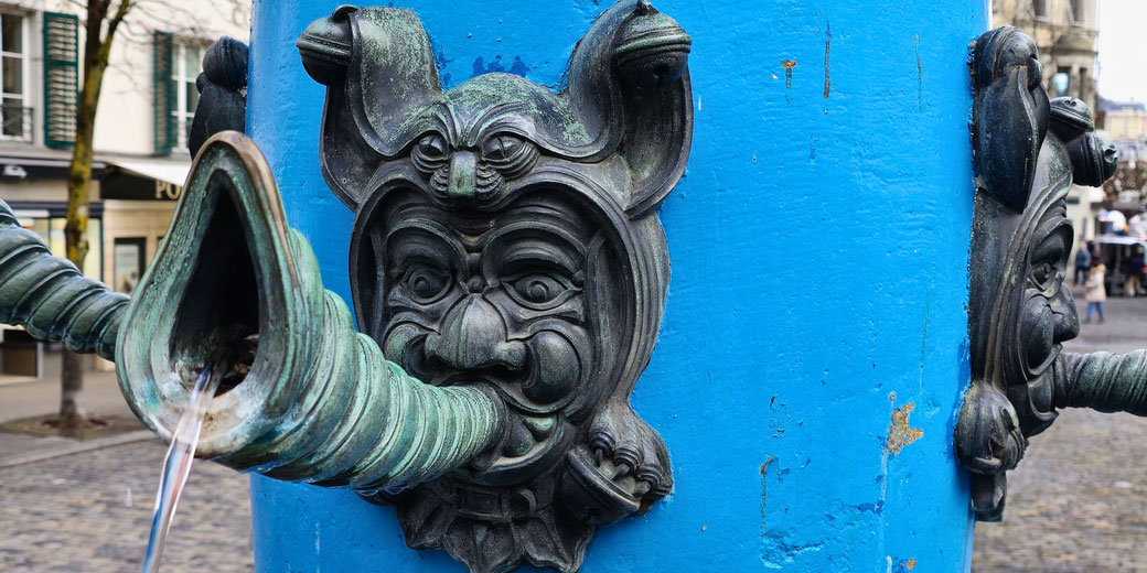 A whimsical bronze fountain with a grotesque face, featuring exaggerated features and a horn-shaped spout flowing with water, set against a bright blue background in an outdoor setting.