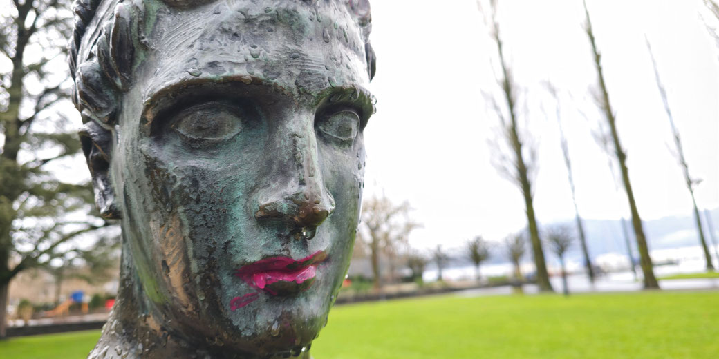 A close-up of a bronze statue's face with a green patina, featuring raindrops and red graffiti on the lips. The background includes a grassy park and blurred trees under an overcast sky.