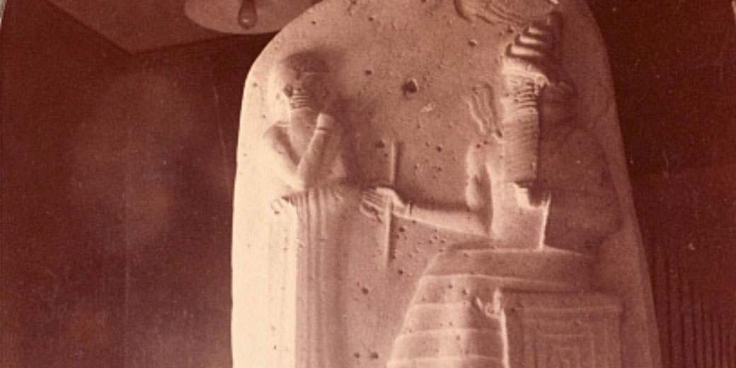 A carved basalt stele shows a seated deity or king handing authority to a standing figure in traditional Mesopotamian dress.