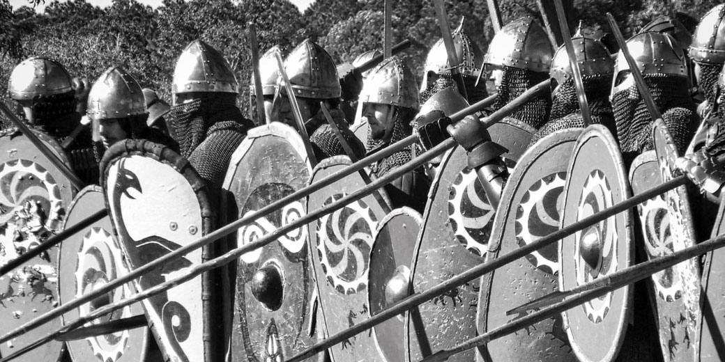 A group of medieval soldiers in chainmail and helmets stand in formation, holding large, round shields with spiral designs.