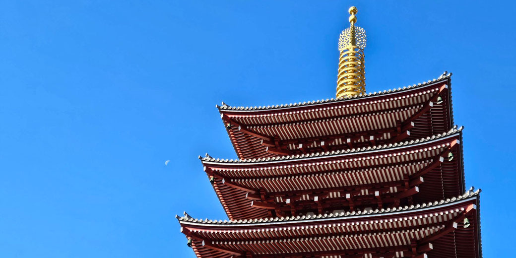 The top tiers of a traditional Japanese pagoda with a golden finial against a clear blue sky. A small crescent moon is visible in the background, adding to the serene composition.