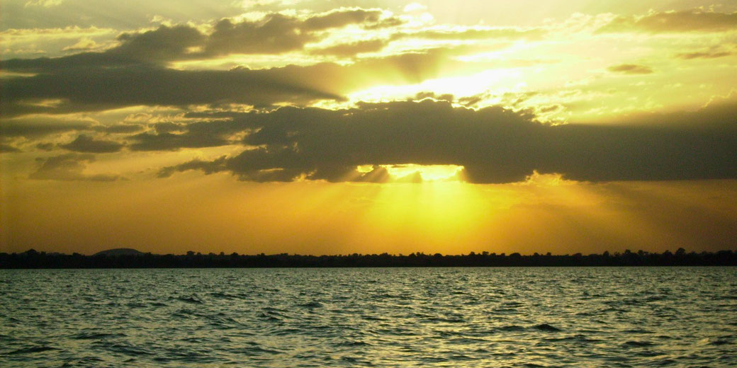 Golden sunset over a body of water with silhouetted treeline and sun rays breaking through dark clouds.