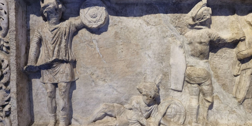 Ancient relief depicting soldiers in helmets and short tunics, one standing with a shield, another fallen on the ground, and a third figure facing away.
