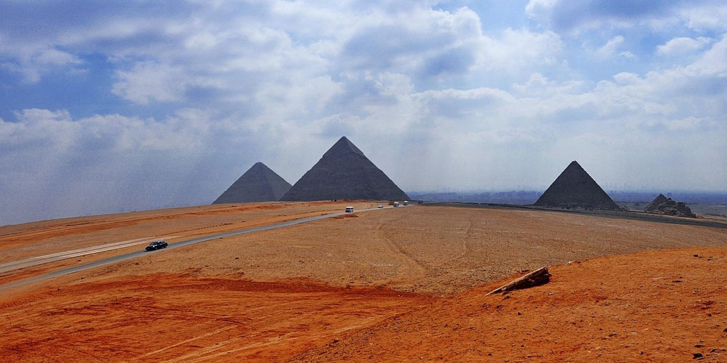 Three pyramids rise from a desert plateau beside a paved road under thick clouds and bright daylight.