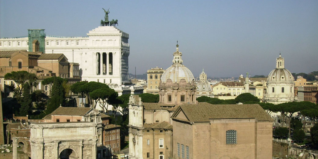 Panoramic view of central Rome featuring historic domes, ancient ruins, and the towering white Vittoriano monument with statue-topped columns.