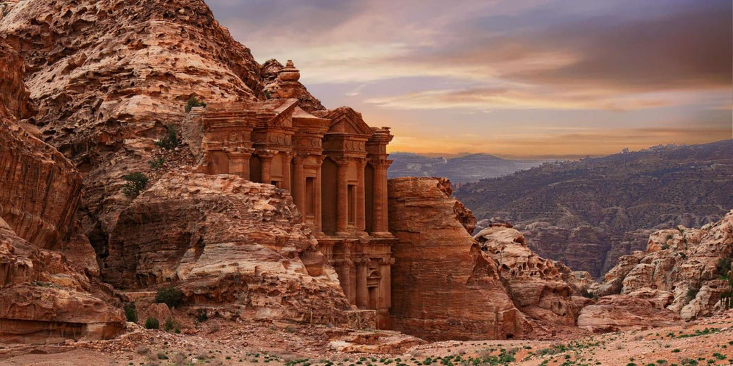 Ancient rock-carved facade set into a reddish cliff, with columns and detailed stonework, surrounded by rugged desert hills under a warm sunset sky.