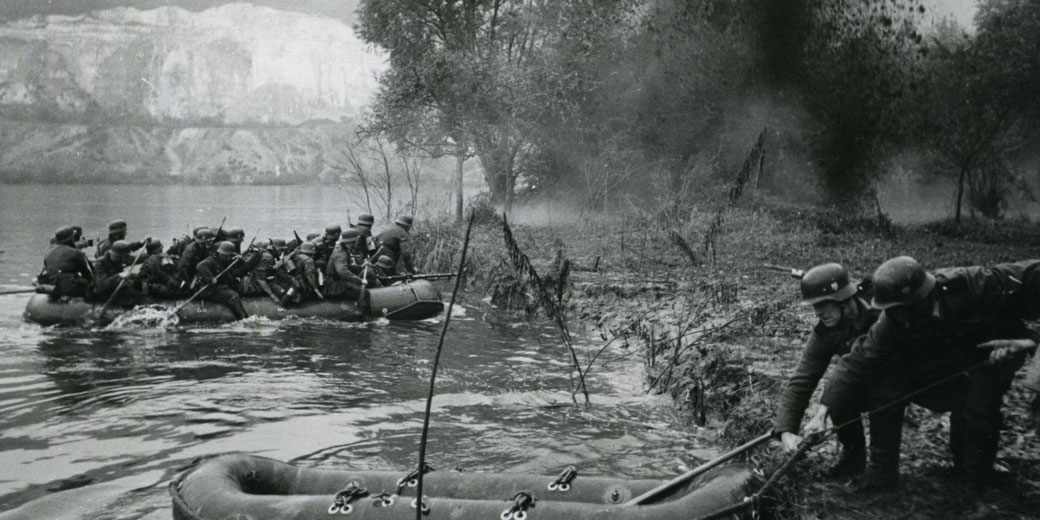 Black and white scene of soldiers crossing a river in an inflatable boat while others pull it ashore, surrounded by trees and mist, suggesting a wartime operation.