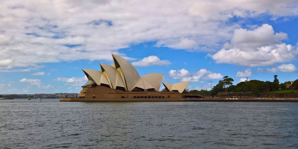 The Sydney Opera House with its iconic sail-like roofs stands by the harbor under a partly cloudy sky.